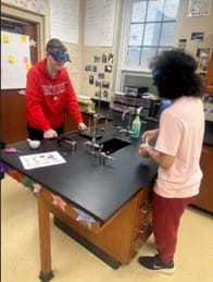 A photo of two students facing each other who appear to be chatting while working in a science lab. One is standing in front of a Bunsen Burner and ring stand setup with a beaker with a yellow solid inside, while the other is using a mortar and pestle. A photo of two students facing each other who appear to be chatting while working in a science lab. One is standing in front of a Bunsen Burner and ring stand setup with a beaker with a yellow solid inside, while the other is using a mortar and pestle.