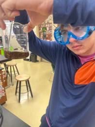 A photo of a student wearing blue goggles and holding up a beaker containing a thick brown fluid to inspect while he stirs. A photo of a student wearing blue goggles and holding up a beaker containing a thick brown fluid to inspect while he stirs.