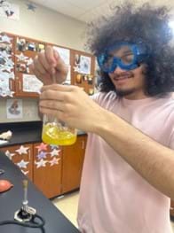 A photo of a student wearing blue goggles who is holding up a beaker containing a thick brown fluid and inspecting it while he stirs. A photo of a student wearing blue goggles who is holding up a beaker containing a thick brown fluid and inspecting it while he stirs.