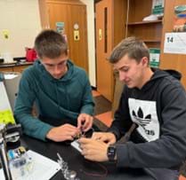 A photo showing two high school students using a breadboard, wires, and other electrical components to build an integrated logic circuit. A photo showing two high school students using a breadboard, wires, and other electrical components to build an integrated logic circuit.