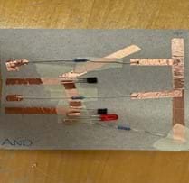 A photo of an AND gate built on a piece of cardboard from a cereal box cut to the size of a notecard. The gate is built using copper tape, three resistors, two transistors and an LED. There is also masking tape used to separate layers of the copper tape to prevent short circuits. There are two places in the circuit where gaps are left between pieces of copper tape to create the switches in their open states. A photo of an AND gate built on a piece of cardboard from a cereal box cut to the size of a notecard. The gate is built using copper tape, three resistors, two transistors and an LED. There is also masking tape used to separate layers of the copper tape to prevent short circuits. There are two places in the circuit where gaps are left between pieces of copper tape to create the switches in their open states.