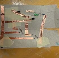 A photo of an OR gate built on a piece of cardboard from a cereal box cut to the size of a notecard. The gate is built using copper tape, three resistors, two transistors and an LED. There is also masking tape used to separate layers of the copper tape to prevent short circuits. There are two places in the circuit where gaps are left between pieces of copper tape to create the switches in their open states. A photo of an OR gate built on a piece of cardboard from a cereal box cut to the size of a notecard. The gate is built using copper tape, three resistors, two transistors and an LED. There is also masking tape used to separate layers of the copper tape to prevent short circuits. There are two places in the circuit where gaps are left between pieces of copper tape to create the switches in their open states.