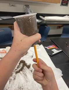 A photo showing a student using a pencil to create holes in a plastic cup to filter potting soil.