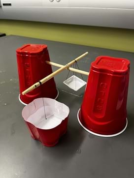 A photo showing the balance for the Method 1 activity. It is a balance beam made from plastic cups, wooden dowels, thread, needle, and weigh basket. A photo showing the balance for the Method 1 activity. It is a balance beam made from plastic cups, wooden dowels, thread, needle, and weigh basket.