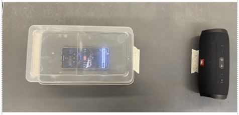 A smartphone displaying a decibel app inside a clear plastic container, next to a black JBL Bluetooth speaker, set up for a sound experiment. A smartphone displaying a decibel app inside a clear plastic container, next to a black JBL Bluetooth speaker, set up for a sound experiment.