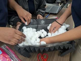 A photo of three people's hands adding cotton balls to a container lined with black sound-dampening foam. A photo of three people's hands adding cotton balls to a container lined with black sound-dampening foam.