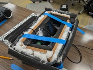 A photo of an open box filled with layers of sound-dampening materials, including foam, cotton, and cork, with blue tape holding them in place. A photo of an open box filled with layers of sound-dampening materials, including foam, cotton, and cork, with blue tape holding them in place.
