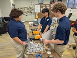 A photo of three students working at a table, examining materials for the activity, with one holding a white board and another student manipulating items on the table. A photo of three students working at a table, examining materials for the activity, with one holding a white board and another student manipulating items on the table.