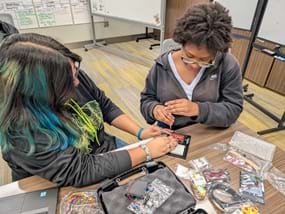A photo of a plastic container filled with dark insulation material sitting on a table, with a clear lid taped shut. A cable runs out of the container to a cardboard piece, next to a reflective, patterned material. A photo of a plastic container filled with dark insulation material sitting on a table, with a clear lid taped shut. A cable runs out of the container to a cardboard piece, next to a reflective, patterned material.