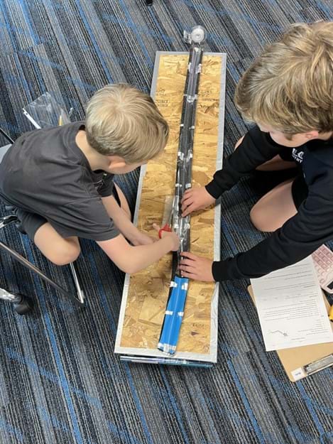 Two students adding plastic material to a foam tube. One student is cutting plastic using the foam tube as a size guide, while the other is holding the plastic to keep it stable for cutting. Two students adding plastic material to a foam tube. One student is cutting plastic using the foam tube as a size guide, while the other is holding the plastic to keep it stable for cutting.