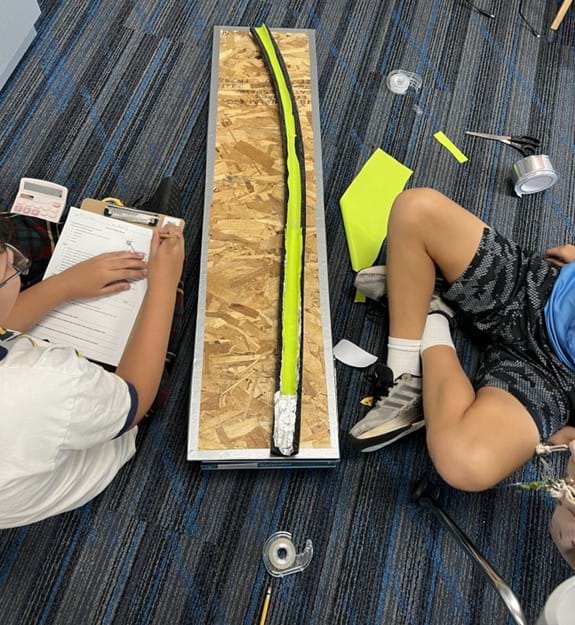 A photo showing two students seated next to a foam tube covered in rubbery material. One student is writing on a data sheet. A photo showing two students seated next to a foam tube covered in rubbery material. One student is writing on a data sheet.
