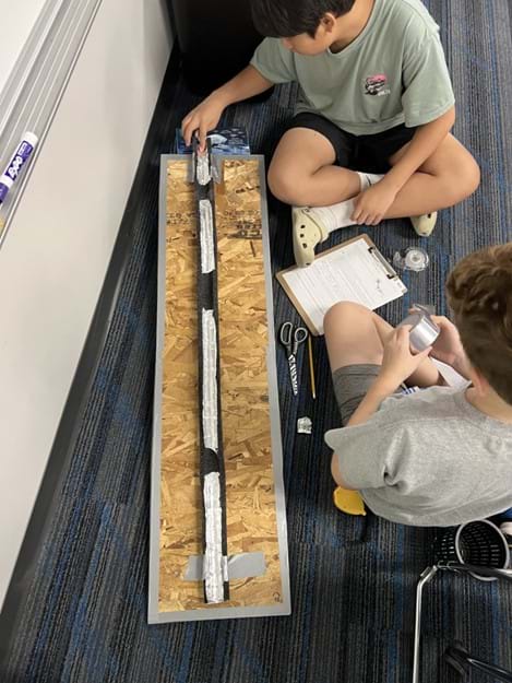 A photo showing two students seated next to a foam tube covered in aluminum material with missing sections of aluminum. One student is about to release a marble into tube, while the other is about to unroll tape to make repairs. A photo showing two students seated next to a foam tube covered in aluminum material with missing sections of aluminum. One student is about to release a marble into tube, while the other is about to unroll tape to make repairs.