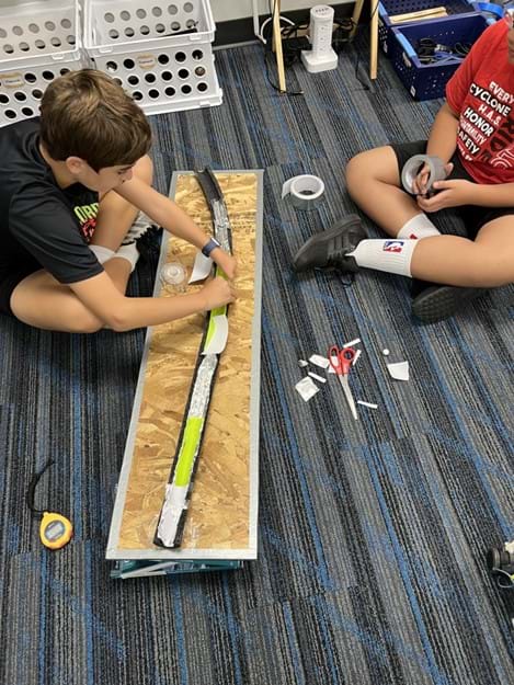 A photo showing two students seated next to a foam tube covered in aluminum and rubbery material. One student is attaching new pieces to the foam to replace damaged pieces, while the other student is about to unroll tape. A photo showing two students seated next to a foam tube covered in aluminum and rubbery material. One student is attaching new pieces to the foam to replace damaged pieces, while the other student is about to unroll tape.