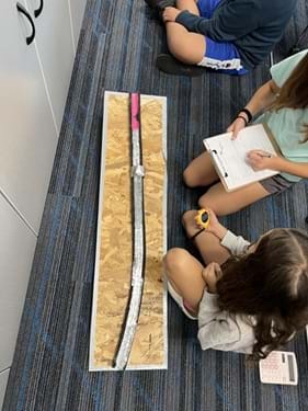 A photo showing two students seated next to a foam tube covered in aluminum and rubbery material. One student is writing on a data sheet, while the other is holding a stopwatch. A photo showing two students seated next to a foam tube covered in aluminum and rubbery material. One student is writing on a data sheet, while the other is holding a stopwatch.