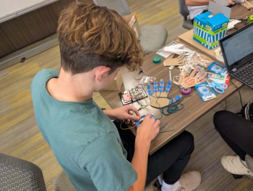 A photo showing a student sitting at a table connecting the wires between his cardboard hand design to the Arduino circuit board. A photo showing a student sitting at a table connecting the wires between his cardboard hand design to the Arduino circuit board.