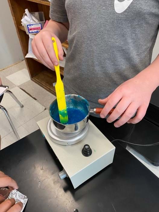 A photo showing a student using a plastic stir stick to mix up the gelatin-based bioplastic recipe. The mixture is blue and is being melted in a pot on a hot plate. A photo showing a student using a plastic stir stick to mix up the gelatin-based bioplastic recipe. The mixture is blue and is being melted in a pot on a hot plate.