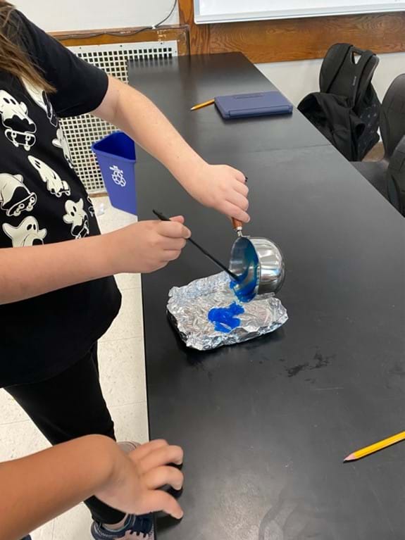 A photo showing a student pouring the blue mixture out of the pot onto a piece of aluminum foil to be flattened and cooled. A photo showing a student pouring the blue mixture out of the pot onto a piece of aluminum foil to be flattened and cooled.