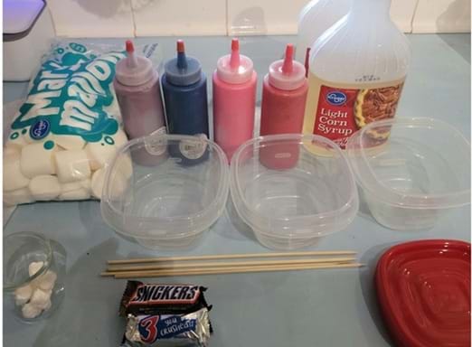 A photo showing materials for a candy-dipping activity. On the left are a bag of large marshmallows and four squeeze bottles containing different colored liquids. Behind the containers is a bottle of light corn syrup. In front are three empty clear plastic containers, two wooden skewers, a small pile of cotton or paper pieces, and a fun-size Snickers candy bar. A photo showing materials for a candy-dipping activity. On the left are a bag of large marshmallows and four squeeze bottles containing different colored liquids. Behind the containers is a bottle of light corn syrup. In front are three empty clear plastic containers, two wooden skewers, a small pile of cotton or paper pieces, and a fun-size Snickers candy bar.
