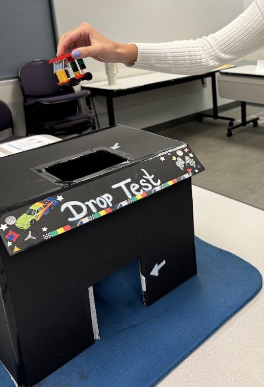 A photo of a student holding a completed plastic brick car over the drop test site. A photo of a student holding a completed plastic brick car over the drop test site.