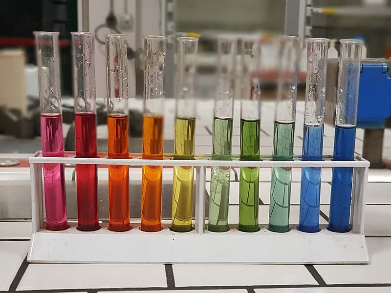 A photo of 10 small glass tubes lined up, representing the pH scale. A rainbow of colors from left to right: pink, red, red-orange, orange, yellow, light green, green, light teal, light blue, blue. 