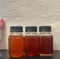 A photo of three small plastic jars filled with a variation of an orange tint of avocado dye. The bottle on the left is slightly lighter in hue than the original base color of orange in the middle jar. The jar on the right is a more brownish color than the original base color of orange in the middle jar. 