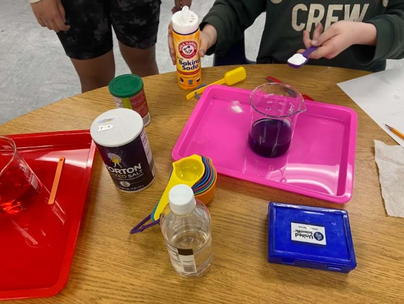 A photo showing two students creating a solution on a table using baking soda, water, salt, food coloring, glass beakers, plastic trays and plastic measuring spoons.