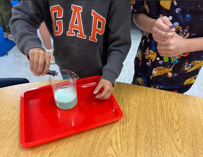 A photo showing two students adding food coloring to a solution in a glass beaker on a plastic tray.