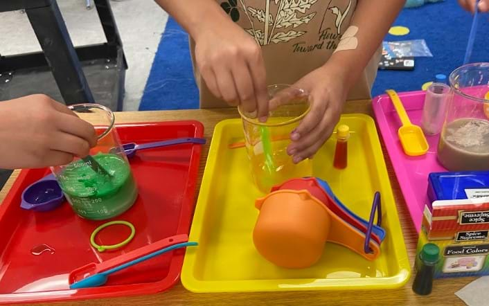 A photo showing two students mixing ingredients in their solutions in glass beakers on plastic trays.