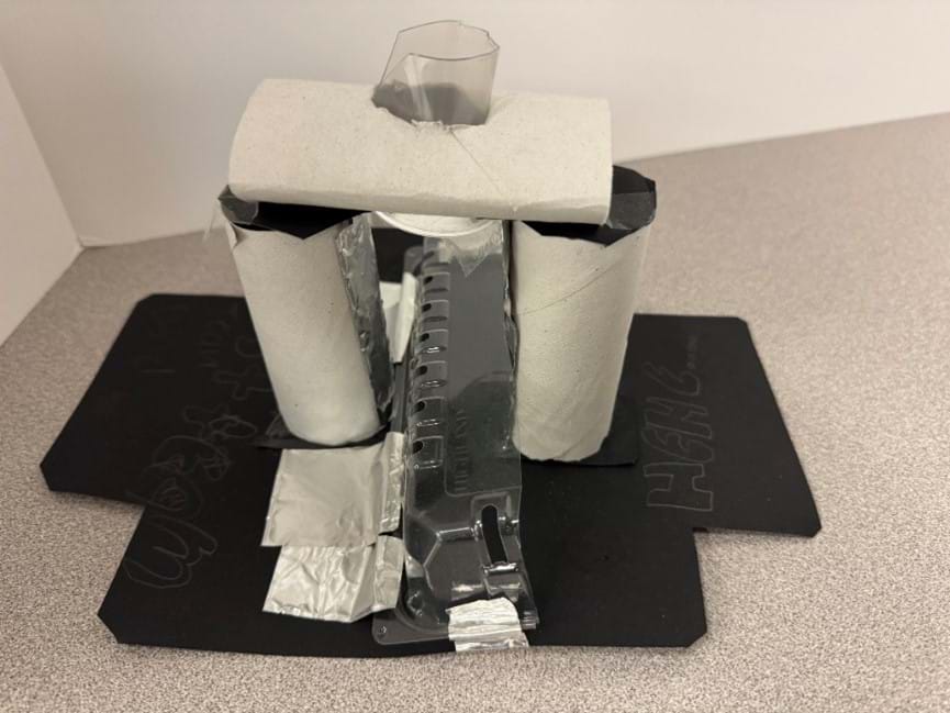 A photo of a DIY microscope model constructed using various household and craft materials. The base of the microscope is made from black cardstock, with visible handwritten text and doodles in white crayon. Two cardboard tubes, from toilet paper rolls, stand vertically as structural supports on each side of the central microscope body. The main body is a repurposed clear plastic container, from food packaging, with silver duct tape used to hold it in place and connect parts. A top horizontal cardboard toilet paper roll bridges the two vertical tubes and supports a transparent plastic piece, which houses the lens or eyepiece. The overall model resembles the general shape of a compound microscope. A photo of a DIY microscope model constructed using various household and craft materials. The base of the microscope is made from black cardstock, with visible handwritten text and doodles in white crayon. Two cardboard tubes, from toilet paper rolls, stand vertically as structural supports on each side of the central microscope body. The main body is a repurposed clear plastic container, from food packaging, with silver duct tape used to hold it in place and connect parts. A top horizontal cardboard toilet paper roll bridges the two vertical tubes and supports a transparent plastic piece, which houses the lens or eyepiece. The overall model resembles the general shape of a compound microscope.