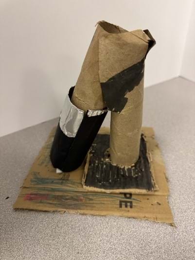 A photo of a DIY microscope model constructed primarily from cardboard materials is displayed on a flat surface. The base consists of a square piece of corrugated cardboard that has been drawn on with various colors, including red, green, and black. Two cardboard tubes are attached vertically to the base—one taller and slightly bent at the top, possibly representing the microscope's eyepiece, and another shorter tube wrapped in black paper and silver duct tape, likely symbolizing the objective lens. Black paint or marker has been used to add details on the tubes and base, giving it a more realistic microscope appearance. A photo of a DIY microscope model constructed primarily from cardboard materials is displayed on a flat surface. The base consists of a square piece of corrugated cardboard that has been drawn on with various colors, including red, green, and black. Two cardboard tubes are attached vertically to the base—one taller and slightly bent at the top, possibly representing the microscope's eyepiece, and another shorter tube wrapped in black paper and silver duct tape, likely symbolizing the objective lens. Black paint or marker has been used to add details on the tubes and base, giving it a more realistic microscope appearance.