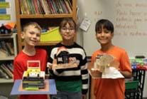 A photo of three students standing, each holding a different model house made from different materials. A photo of three students standing, each holding a different model house made from different materials.