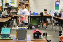 A photo showing a student pointing a leaf blower at three model houses while two students sit behind their desks watching. A photo showing a student pointing a leaf blower at three model houses while two students sit behind their desks watching.